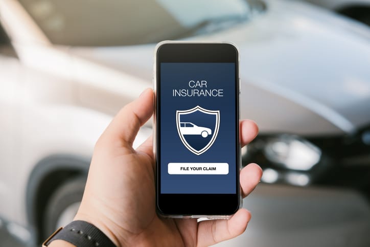 An upclose image of a man holding a cell phone with a vehicle in the background and the phone screen says car insurance on it