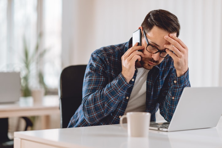 A young man with glasses leans in consternation over the laptop and holds his hand to his forehead while making a serious phone call