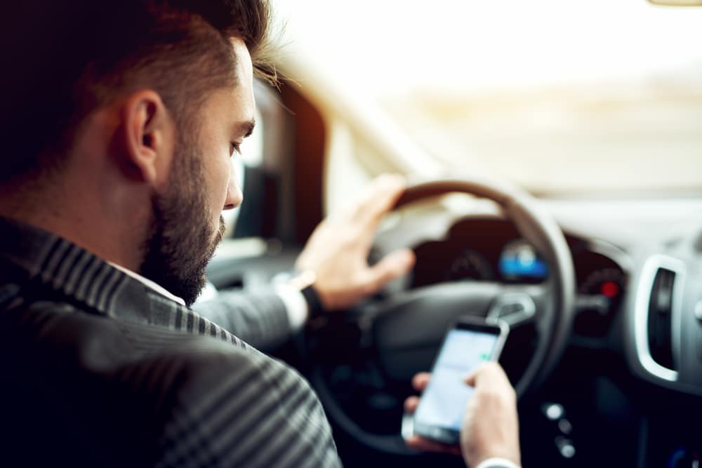 A man behind a steering wheel looks at his phone and texts while holding the wheel with one hand