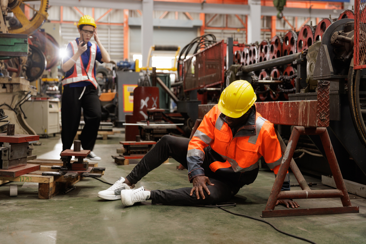 Injured factory worker sitting on the floor near industrial machinery after a workplace accident requiring medical treatment.